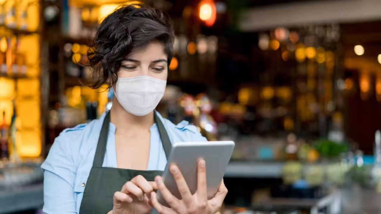 Portrait of a happy waitress wearing a facemask at a restaurant and using a tablet computer during the COVID-19 pandemic